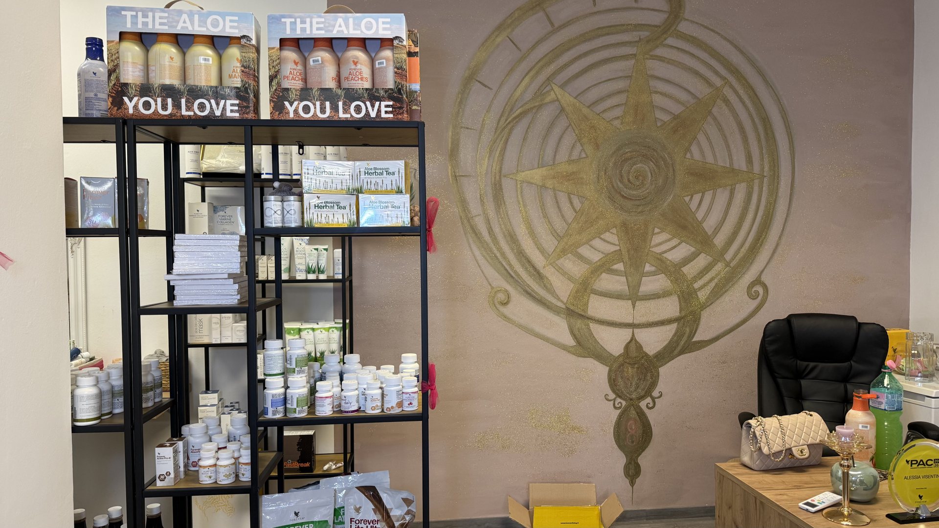 Scaffale nero con confezioni The Aloe You Love, tisane e integratori accanto a una rosa dei venti dorata dipinta a muro