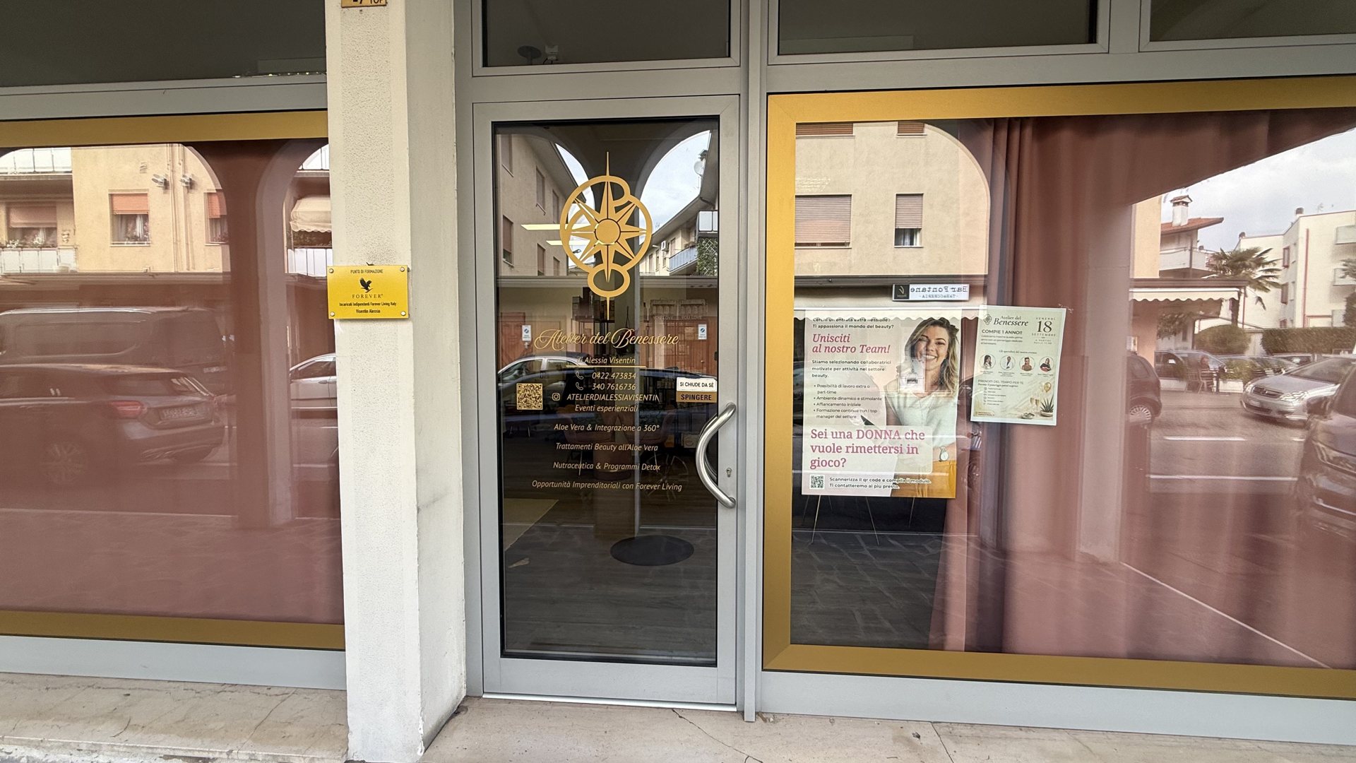 Ingresso dell'Atelier del Benessere con porta a vetri, rosa dei venti dorata, tende rosa e locandina «Unisciti al nostro Team!